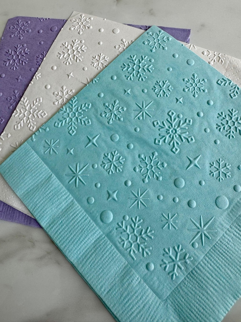 May include: Three decorative paper napkins in shades of purple, white, and blue. Each napkin features an embossed snowflake design with stars and circles. The napkins are ideal for winter-themed events or holiday gatherings.