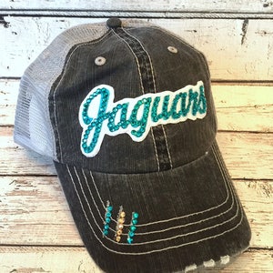 May include: A distressed black baseball cap with a gray mesh back. The word "Jaguars" is spelled out in teal rhinestones. The brim has decorative rhinestones in teal and gold.