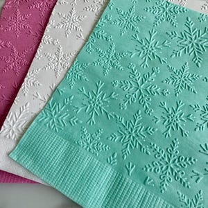 Puede incluir: Primer plano de tres servilletas de papel en tonos rosa, blanco y turquesa. Cada servilleta presenta un patr&oacute;n de copos de nieve en relieve. La servilleta turquesa est&aacute; en primer plano, con un borde texturizado.
