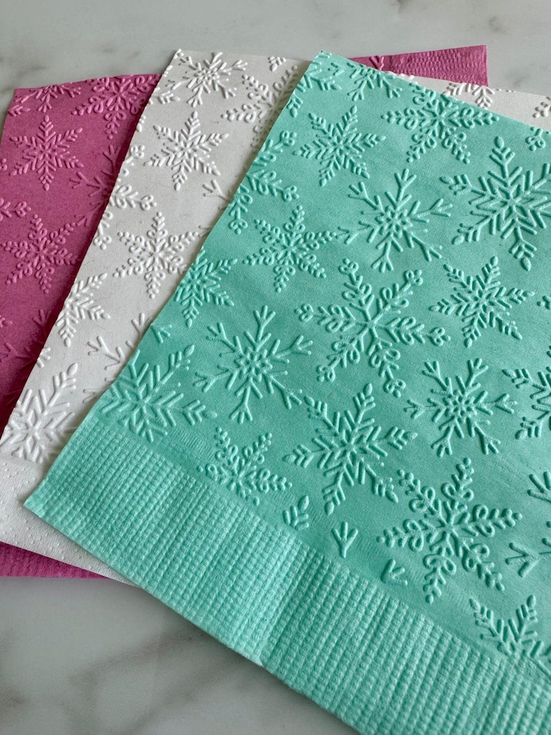 Puede incluir: Primer plano de tres servilletas de papel en tonos rosa, blanco y turquesa. Cada servilleta presenta un patr&oacute;n de copos de nieve en relieve. La servilleta turquesa est&aacute; en primer plano, con un borde texturizado.