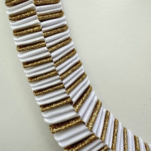 May include: Close-up of a decorative white and gold ribbon. The ribbon is pleated with alternating sections of white fabric and gold glitter. The design is arranged in a curved shape, suggesting it could be used for embellishment or crafting.