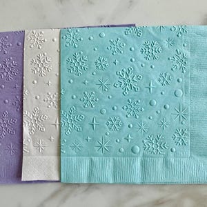 May include: Three decorative paper napkins in shades of purple, white, and blue. Each napkin features an embossed snowflake pattern with stars and dots. The napkins are ideal for winter-themed events or holiday gatherings.