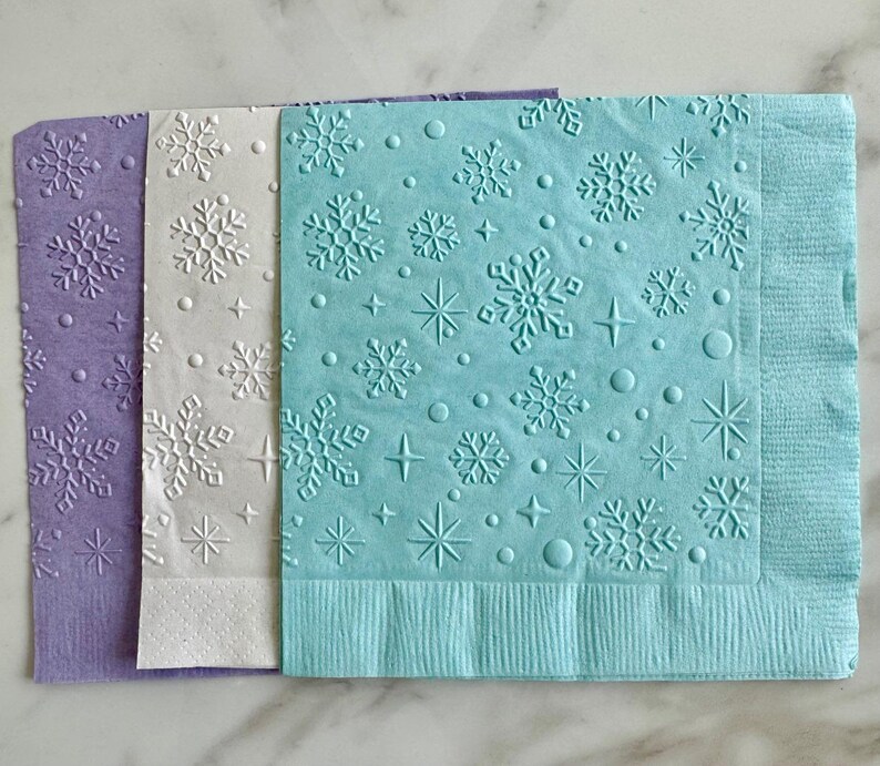 May include: Three decorative paper napkins in shades of purple, white, and blue. Each napkin features an embossed snowflake pattern with stars and dots. The napkins are ideal for winter-themed events or holiday gatherings.