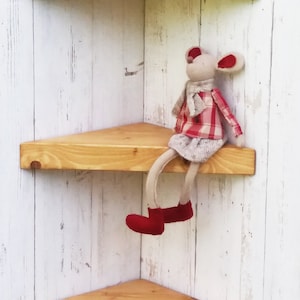 Può includere: Tre mensole d'angolo in legno con finitura in legno naturale. Le mensole sono montate su una parete bianca. Un piccolo giocattolo di peluche a forma di topo è seduto sulla mensola centrale.