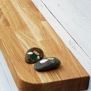 Pu&ograve; includere: Uno scaffale in legno con bordi arrotondati, montato su una parete bianca. Due pietre dipinte con figure fantasiose sono posizionate sullo scaffale.