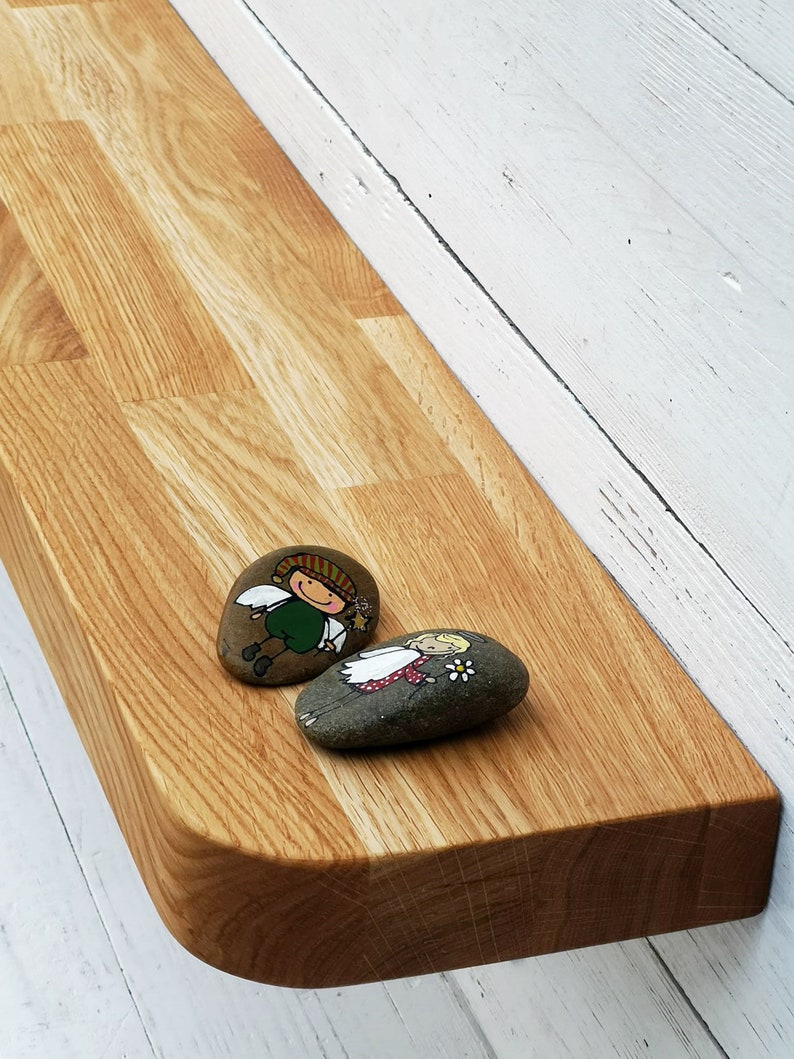 Pu&ograve; includere: Uno scaffale in legno con bordi arrotondati, montato su una parete bianca. Due pietre dipinte con figure fantasiose sono posizionate sullo scaffale.