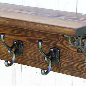 Può includere: Uno scaffale in legno con tre ganci in metallo e staffe metalliche decorative. Lo scaffale è tinto di un colore marrone scuro e i ganci e le staffe sono di colore argento.