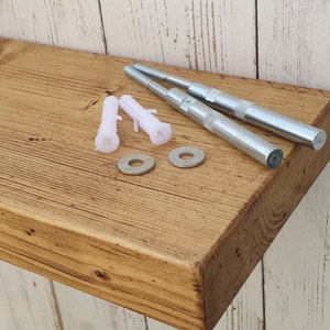 May include: A rustic wooden shelf with a natural finish. The shelf is made of reclaimed wood and has a distressed look. It comes with two metal brackets and four plastic anchors for mounting.