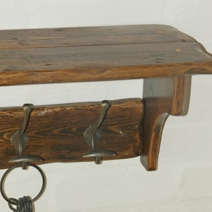 May include: A rustic wooden shelf with three metal hooks, perfect for hanging coats, hats, or bags. The shelf has a dark brown stain and a natural wood finish. The hooks are made of metal and have a brushed finish.