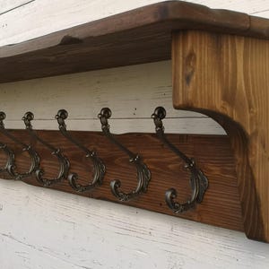 Può includere: Un appendiabiti in legno rustico con uno scaffale e sei ganci in metallo decorativi. L'appendiabiti è tinto di un colore marrone scuro e ha una finitura invecchiata.