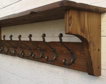 May include: A rustic wooden coat rack with a shelf and six ornate metal hooks. The rack is stained a dark brown color and has a distressed finish.