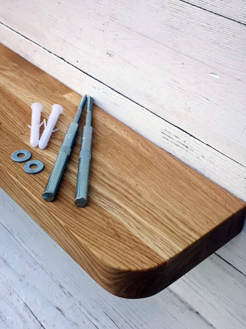 May include: A wooden shelf with rounded edges, made of light brown oak wood. The shelf is attached to a white wall with two silver screws and two white plastic anchors.