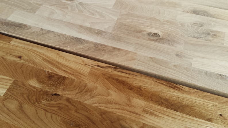 May include: Two pieces of wood flooring, one stained a darker brown and the other left in its natural light wood color. The wood grain is visible in both pieces.