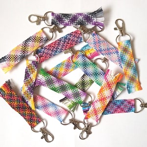 May include: A collection of rectangular woven keychains featuring various multicolored geometric patterns. Each keychain is crafted from vibrant thread and attached to a silver-toned metal lobster clasp, displaying a mix of bright hues.