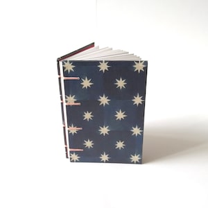 May include: A small notebook with a navy blue cover adorned with cream-coloured star shapes. The binding is visible, stitched with pink thread. The pages are white, and the cover edges are red.