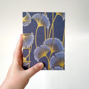 Op de afbeelding: Een rechthoekig notitieboek met een marineblauwe achtergrond en een goud- en blauw ginkgo-bladpatroon. Het notitieboek wordt door een hand tegen een witte achtergrond gehouden.