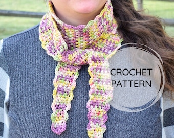 May include: A crochet scarf pattern featuring a scalloped edge and a mix of purple, yellow, and green yarn. The pattern is shown on a model wearing a grey sweater.