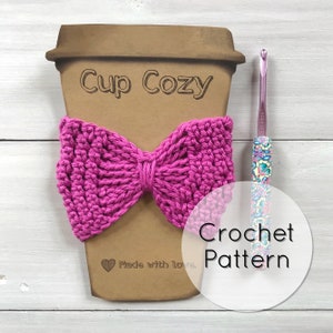 Może przedstawiać: Różowy, dziergany na szydełku, ozdobny uchwyt na kubek w kształcie kokardki z napisem "Cup Cozy" i "Made with Love" na brązowym papierowym kubku. Wokół kubka znajduje się napis "Crochet Pattern" w kółku. Na zdjęciu widoczny jest również szydełkowy hak.