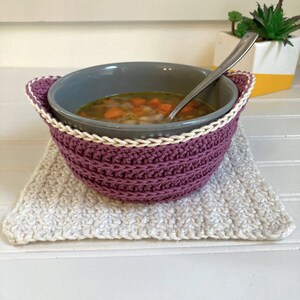 May include: A purple crocheted bowl cozy with a white crocheted trim, holding a gray bowl filled with soup. The bowl cozy is sitting on a white crocheted coaster.