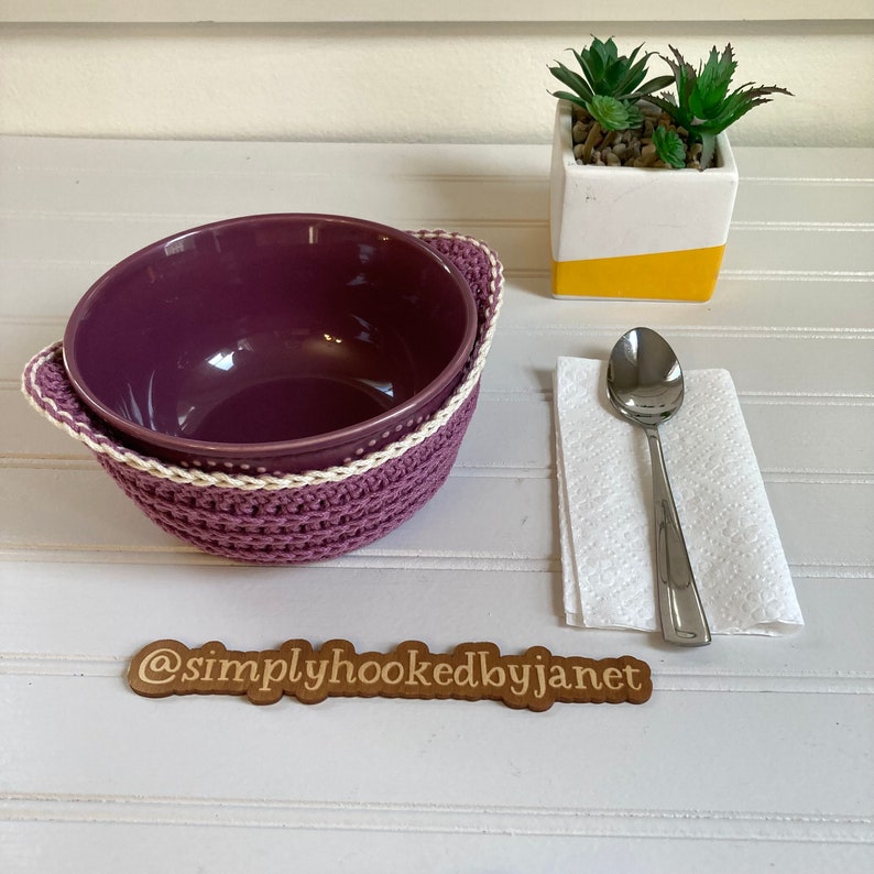 May include: Purple ceramic bowl with a white crocheted bowl cozy. The cozy has a white trim. A spoon and a white napkin are on a white wooden table. A small potted plant is in the background. The image includes the text "@simplyhookedbyjanet".