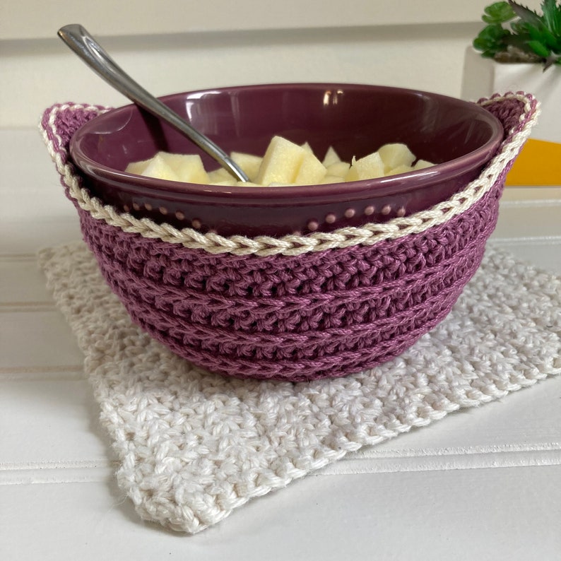 May include: A purple ceramic bowl with a white crocheted bowl cozy. The bowl is filled with chopped apples and a silver spoon is resting on the rim. The bowl is sitting on a white crocheted square.