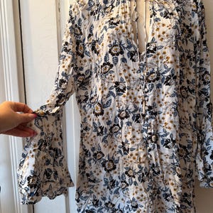 Puede incluir: Blusa blanca con estampado floral, mangas largas y cuello con volantes. La blusa presenta una abertura de ojo de cerradura en el pecho y un delicado estampado floral en negro, azul y dorado.