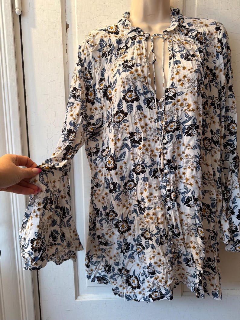 Puede incluir: Blusa blanca con estampado floral, mangas largas y cuello con volantes. La blusa presenta una abertura de ojo de cerradura en el pecho y un delicado estampado floral en negro, azul y dorado.