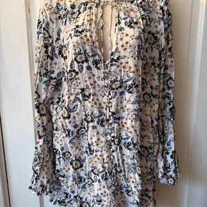 Puede incluir: Una t&uacute;nica blanca de manga larga con un estampado floral. El estampado incluye flores y hojas negras, azules y doradas. La t&uacute;nica tiene un cuello con volantes y un escote anudado.