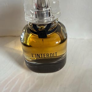 Puede incluir: Un frasco de perfume con una tapa transparente y angular y un atomizador plateado. La botella está llena de un líquido dorado y tiene una cinta negra alrededor del cuello. Las palabras "L'INTERDIT" están impresas en la parte delantera.