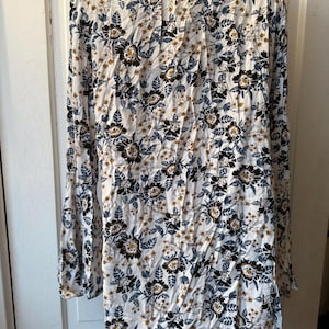 Puede incluir: Vestido de manga larga con estampado floral. El vestido presenta un fondo blanco con dise&ntilde;os florales negros, azules y amarillos. Tiene un cuello alto y mangas acampanadas, creando un estilo bohemio.