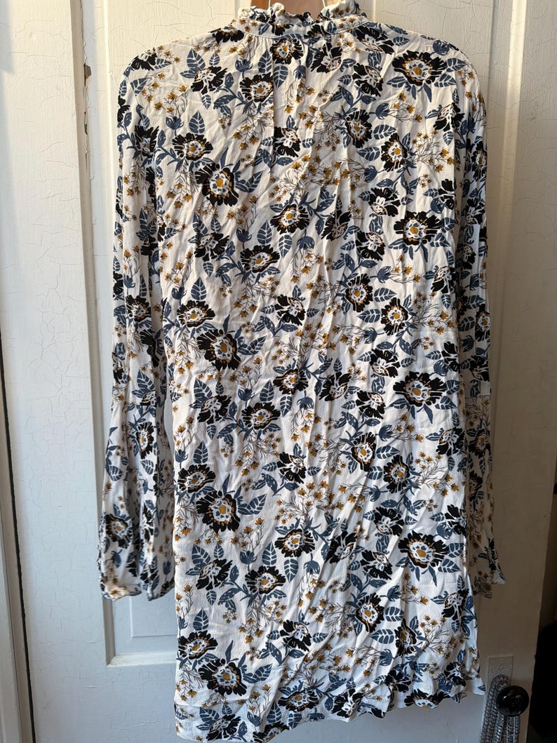 Puede incluir: Vestido de manga larga con estampado floral. El vestido presenta un fondo blanco con dise&ntilde;os florales negros, azules y amarillos. Tiene un cuello alto y mangas acampanadas, creando un estilo bohemio.