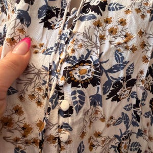 Puede incluir: Primer plano de una blusa floral blanca con botones en la parte delantera. La tela presenta un estampado de flores y hojas negras, azules y doradas. La blusa tiene un escote en V y un detalle de lazo. Los botones son blancos.