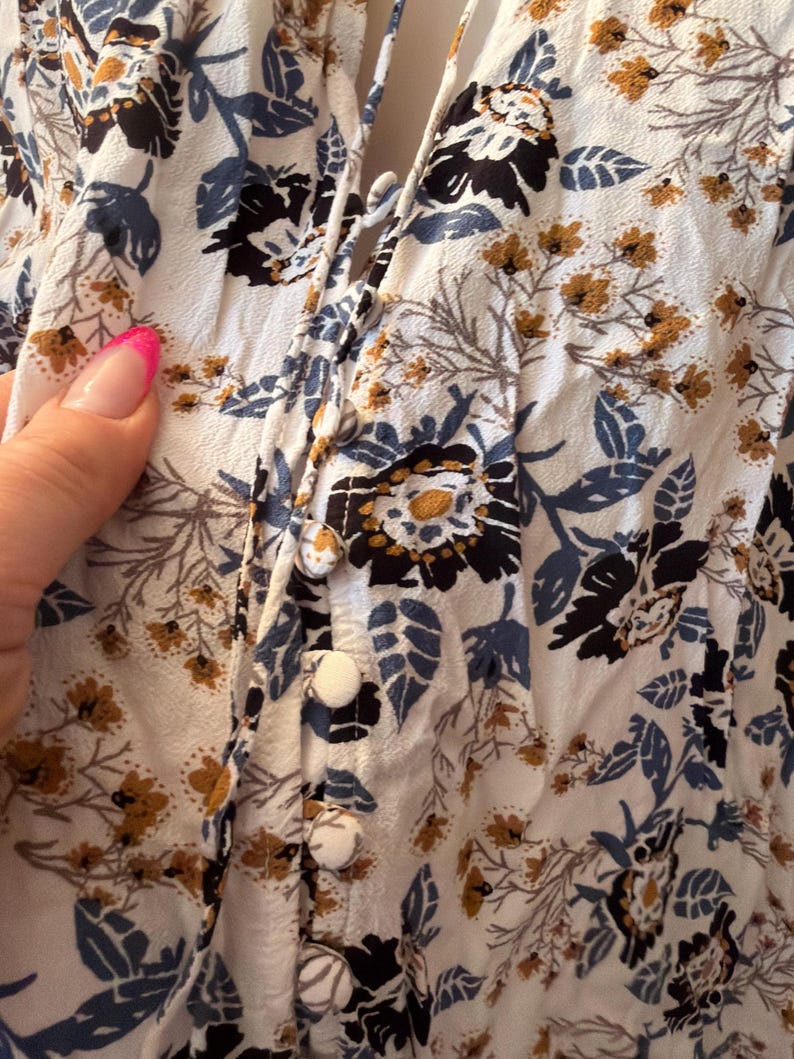 Puede incluir: Primer plano de una blusa floral blanca con botones en la parte delantera. La tela presenta un estampado de flores y hojas negras, azules y doradas. La blusa tiene un escote en V y un detalle de lazo. Los botones son blancos.