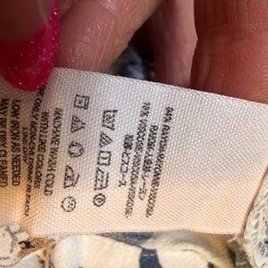Puede incluir: Una etiqueta de tela blanca con instrucciones de cuidado y detalles de la composici&oacute;n de la tela. La etiqueta incluye texto en ingl&eacute;s, espa&ntilde;ol y japon&eacute;s, con s&iacute;mbolos de lavado. La tela es 64% ray&oacute;n, 16% viscosa.