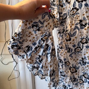 Puede incluir: Blusa blanca con estampado floral en negro, azul y dorado. La blusa tiene mangas con volantes y un corte holgado, ideal para un look casual.