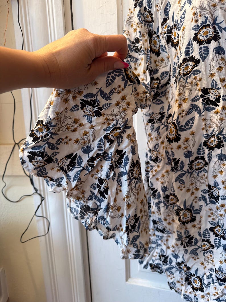Puede incluir: Blusa blanca con estampado floral en negro, azul y dorado. La blusa tiene mangas con volantes y un corte holgado, ideal para un look casual.