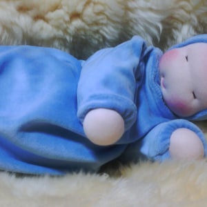 May include: A soft, blue baby doll with a white hat and a rosy complexion. The doll is lying on a white fluffy surface.
