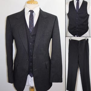 May include: A charcoal gray three-piece suit with subtle pinstripes. The suit includes a jacket, vest, and trousers. The jacket has a two-button closure, and the vest has a button-down front. A white dress shirt and patterned tie complete the ensemble.
