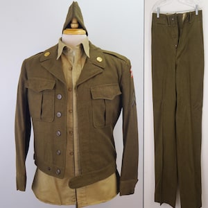 May include: Olive green military uniform consisting of a jacket, shirt, pants, and cap. The jacket has two chest pockets, button closures, and gold-colored buttons. The shirt is tan, and the pants are olive green. The cap is olive green.