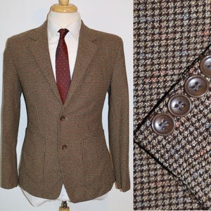 May include: A brown, beige, and red houndstooth patterned blazer with two button closures. The blazer has flap pockets and a brown button detail on the sleeve.