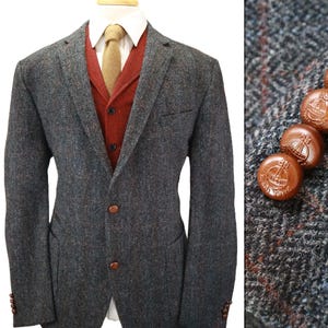 May include: A charcoal gray herringbone tweed blazer with a rust-colored vest, a white collared shirt, and a tan necktie. The blazer features brown buttons embossed with a globe and cross emblem, visible in a close-up detail on the right side.