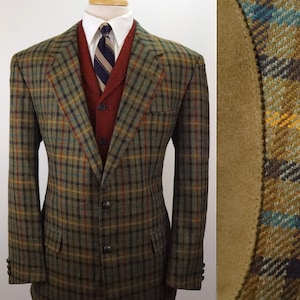 May include: A plaid wool blazer in shades of olive green, brown, and blue with tan suede elbow patches, worn over a rust-colored knit vest, a white collared shirt, and a navy, tan, and white striped necktie on a mannequin torso display.
