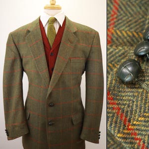 May include: An olive green herringbone blazer with a red windowpane check pattern, featuring two front buttons and two flap pockets. Underneath is a white collared shirt, a red knit cardigan, and a green tie. The sleeve cuffs have small dark buttons.