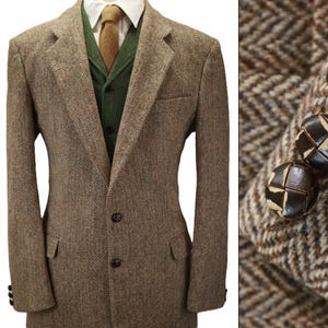 May include: A brown and tan herringbone tweed blazer featuring notched lapels, two front buttons, and two flap pockets. The blazer is worn over a green vest, a white collared shirt, and a tan tie. The sleeve cuffs feature dark brown leather buttons.