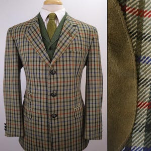 May include: A three-button blazer featuring a tan, brown, and green plaid pattern with red and blue accents. The blazer is worn over a white collared shirt and a green textured vest. A close-up detail shows a brown suede elbow patch on the sleeve.