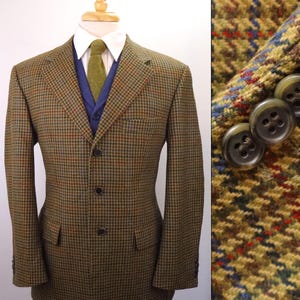 May include: A brown, green, and blue tweed blazer featuring a houndstooth pattern, three front buttons, and two flap pockets. The blazer is worn over a white collared shirt, a blue vest, and a textured olive green necktie. The sleeve cuffs feature three dark buttons.
