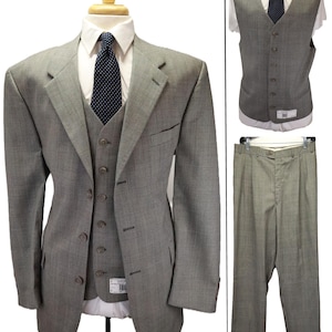 May include: A three-piece suit in a light gray fabric featuring a blazer, a five-button vest, and pleated trousers. The ensemble is paired with a white collared shirt and a dark navy blue necktie with a white polka dot pattern.