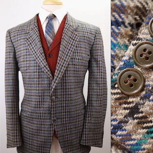 May include: A blazer featuring a multi-colored plaid pattern in shades of blue, brown, and black, worn over a rust-colored vest, a white collared shirt, and a blue plaid tie. The sleeve cuff displays three dark brown buttons in a vertical row.