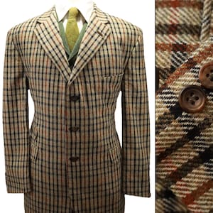May include: A tan, brown, and black plaid blazer featuring a three-button front closure, notched lapels, and two flap pockets. The blazer is worn over a white collared shirt and a textured olive green necktie, with brown buttons on the sleeve cuffs.
