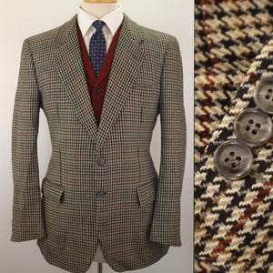 May include: A brown, black, and cream houndstooth blazer with two front buttons and two flap pockets. The blazer is worn over a dark red V-neck sweater, a white collared shirt, and a dark blue patterned tie. The sleeve features three round buttons.